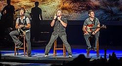 Three men performing music on a stage. One is singing and the others are playing guitars. All three are sitting on stools.