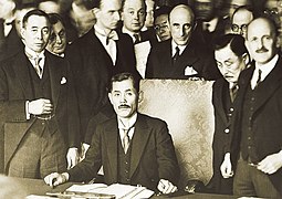 photo d'un homme en costume assis sur une chaise de grande taille en train de signer un document. Autour de lui, d'autres hommes assistent à la scène.