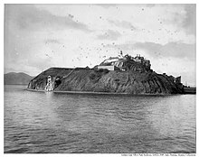 Photo en noir et blanc d'une île rocheuse surmontées de bâtiments. beaucoup d'oiseaux volent au-dessus de l'île.