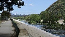 Riverwalk with low wall and natural stream, skyscrapers in far distance