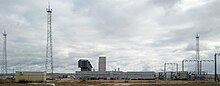 Des installations industrielles comportant plusieurs tours et structures métalliques, à droite et à gauche de l'image et des bâtiments, devant un ciel nuageux.