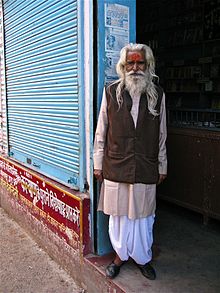 Vieil homme à Rishikesh sur le pas de sa boutique, portant le costume traditionnel : pantalon dhotî blanc, veste achkan brune sur kurta de coton beige saumon à patte boutonnée