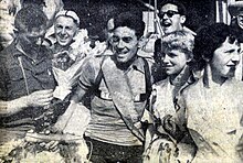 Photo en noir et blanc d'un homme en tenue de cycliste avec une femme blonde à sa gauche. D'autres personnes sont en arrière plan de la photo.