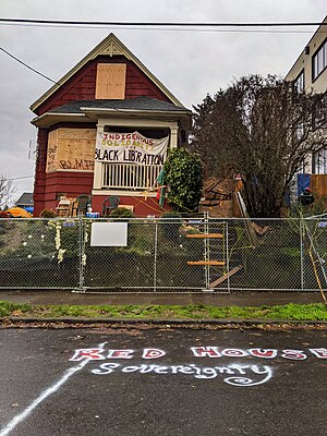 Daytime photograph of a red house, with plywood over the windows. A wooden fence in front of the home has a "Private Property" sign on it.