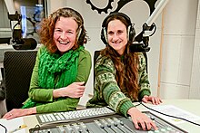 Zwei Frauen am Schaltpult eines Radios, beide tragen Kopfhörer, vor ihnen ein Radiomikrofon