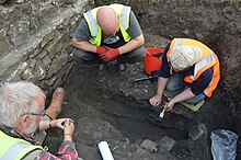 Three individuals crouch in a trench in the process of excavating an archaeological feature.
