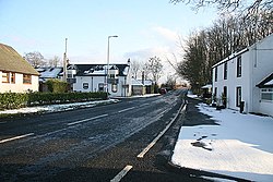 Single carriageway road stretching into distance with houses either side in light snow covered ground on a sunny day with a pale blue sky