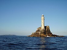 Phare sur un rocher isolé en mer. En arrière-plan, on aperçoit la côte.