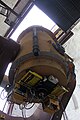 A view looking up along the telescope tube. In the background, the open observing slit in the dome shows an overexposed sky. In the foreground, some electronic devices are mounted on the bottom end of the telescope tube, with messy cables. There are two orange boxes, two yellow racks of some kind, and a few other devices. On the side of the telescope tube directly in front of the camera is a narrow black cylinder; to its right, a set of weight plates is bolted to the telescope tube.