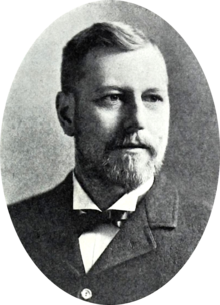 A black and white portrait of a bearded man, wearing a suit with a bowtie