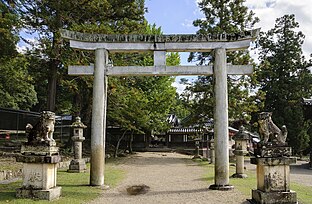 Vista de la entrada norte del recinto del santuario formada por un torii de piedra gris flanqueada por dos leones del mismo material, rodeados de bosque de pinos y cedros.