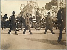 Several men walk down the road. Roosevelt sits in a horse-drawn carriage. A large crowd watches in the distance.