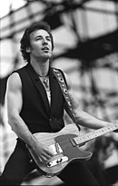 A black and white photo of a caucasian man dressed in a sleeveless blazer and muscle shirt performing with a guitar on stage.