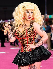 Photograph of someone on a pink runway, wearing an outfit which is black on the bottom half and has an animal print on the top half; she has multiple tattoos and is holding a small stuffed dog in one of her hands