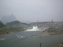 A grey dam in the distance spilling water from its center. Mountains are in the background.