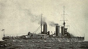 Black and white photograph of a grey warship, a high forward mast above its conning tower, and smoke rising from 2 of its funnel stacks