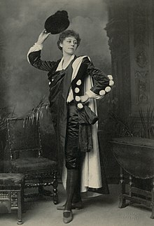 Young woman with short curly hair standing in flamboyant male clothing, with cape in indoor setting