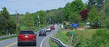 A group of cars, seen from behind, traveling down a slightly curved two-lane paved road with a double-yellow stripe in the middle through an area of small fields and trees, in summertime. There are metal guardrails along the sides, longer on the left than the right, and an entranceway on the right further down with a stone and wood sign. Next to the road in the foreground of the image is a small white on blue metal sign promoting the adopt-a-highway program, with a smaller white on green metal sign with numbers and letters further down the signpost.