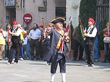 Photo shows a man in early 1800s garb with a short rifle. He wears a blue coat and trousers with a yellow waistcoat, a black tricorne hat with yellow trim, and white knee stockings.