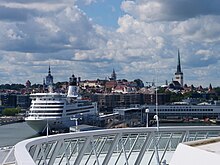 Farbfotografie über eine moderne Brücke zum Kreuzfahrtschiff an der Anlegestelle. Im Hintergrund Häuser und Kirchtürme.