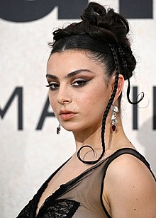 A woman, poised in a black dress with some sheer fabric, has two braids visible coming from her black hair, has eye makeup and muted lipstick, and the facial expression of a model.