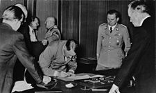 Hitler při podepisování Mnichovské dohody. Vzadu v pozadí stojí Hermann Göring, který rozmlouvá s Mussolinim. Vedle Hitlera stojí Schaub a muž opřený o stůl je Joachim von Ribbentrop