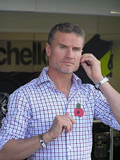 Upper body of a man in his late thirties with a shaved, greying beard, wearing a T-shirt with light blue and dark red lines. He is holding his left arm towards his face and is sporting a poppy on the left of his T-shirt.