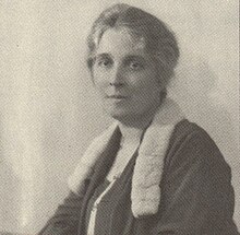 A seated woman, gray hair pinned up, in a black dress with fur collar before a plain white background