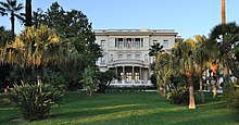 Vue d'un jardin avec palmiers au fond duquel une bâtisse blanche imposante à toit plat et entrée à colonnes se détache.