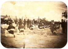 An old photograph showing a procession passing between lines of soldiers with tents in the background