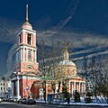 Seitliche Farbfotografie einer rosa Kirche mit einem Turm aus Säulen und einer goldenen Stabspitze auf der linken Seite. Über dem Säuleneingang befindet sich ein Giebelfeld. Rechts befindet sich ein weiterer Säuleneingang mit Giebelfeld und einem Kuppeldach sowie einer goldenen Spitze. Ein flaches Zwischengebäude verbindet beide Gebäudeteile.