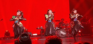 Three women wearing similar costumes consisting of a metallic top and dark skirt, performing on stage lit in bright red