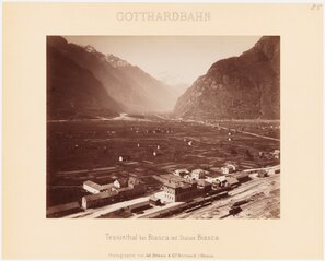 Diese Schwarz-Weiss-Aufnahme von ca. 1870 zeigt den Bahnhof Biasca mit den damals noch sehr wenigen umgebenden Bauten. Es handelt sich mehrheitlich um Holzschuppen und drei Anordnungen fest gemauerter Häuser. Im Vordergrund sind Wagen, eine Lokomotive und Geleise zu sehen. Im Mittelgrund erstreckt sich der landwirtschaftlich genutzte Talboden. Im Hintergrund erheben sich die umliegenden Berge.