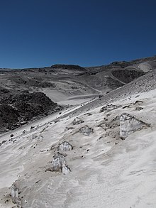 Blue-ish ice emerges from underneath grey sand on a mountain slope