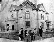 Black and white photo of the Smoot family in front of their home in Provo, Utah. The family is dressed in typical turn-of-the-century attire. Reed and his wife, Allie, are seen with five of their children, including one baby in Allie's arms. A medium-sized dog is sitting in front of the family.
