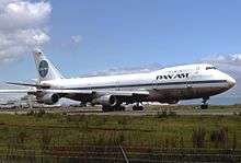 Photo du côté droit d'un avion au sol, légèrement de face, aux couleurs blanches avec une ligne bleue sur le milieu du fuselage, une inscription « Pan Am » à l'avant et le logo circulaire bleu et blanc de la compagnie sur la queue.