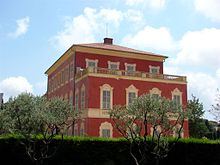 Vue d'un bâtiment en rouge avec façade en trompe-l'œil sous un ciel bleu avec deux arbres et des haies en premier plan.