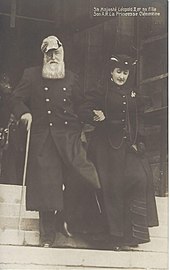 Photo de Léopold II en tenue de lieutenant-général, portant une barbe blanche et coiffé d'un bicorne, s'appuyant de la main droite sur une canne et donnant le bras gauche à sa fille Clémentine, vêtue d'un long manteau sombre et coiffée d'un petit chapeau.Tous deux descendent un escalier.