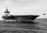 Large aircraft carrier with boxy island superstructure and numerous aircraft on its flight deck and one aircraft being launched.