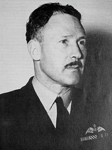 Portrait d'un homme moustachu en uniforme militaire sombre avec des ailes de pilote sur la poche gauche.