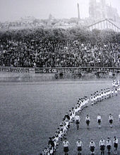 Défilé d'athlètes devant une tribune pleine de spectateurs.