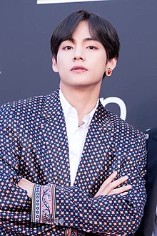 Upper body shot of V posing with arms folded wearing a dark blue, patterned jacket and a large, round gold earring with a red jewelled center on his left ear