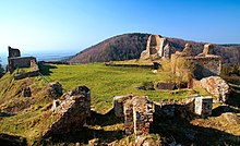 Fotografie sluncem zalitých obvodových zdí zříceniny na vrcholu kopce. V pozadí vedlejší hustě zalesněný vrch, v dáli rovina.