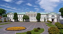 Frontale Farbfotografie eines weißen, zweigeschossigen Gebäudes mit grünem Dach. Im mittleren Obergeschoss bilden Säulen eine Loggia. Vor dem Gebäude ist ein großer Platz mit einem runden Beet mit gelben Blumen. Am Rand sind kleine Rasenflächen und einige junge Bäume.