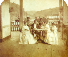 a man and three women seated around a table with two servants standing in the background