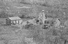 Photo noir et blanc d'un chevalement en bois encadré par deux petits bâtiments maçonnés.