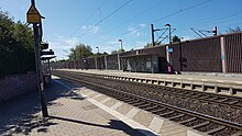 zwei Seitenbahnsteige an einer zweigleisigen elektrifizierten Bahnstrecke, rechts eine Schallschutzwand