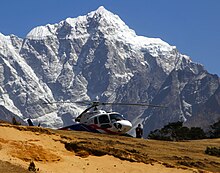 Photographie couleurs en contre-plongée d'un hélicoptère posé sur un sol rocheux. Une montagne couverte de neige se détache nettement en fond.