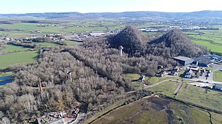 Vue aérienne d'une forêt parsemée de ruines industrielles et bordée par deux grands terrils coniques.