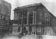 Photographie en noir et blanc, au premier plan, sur la gauche, la devanture et l'enseigne d'un café, à l'arrière plan, sur une place, un bâtiment en pierre imposant de style néo-classique.